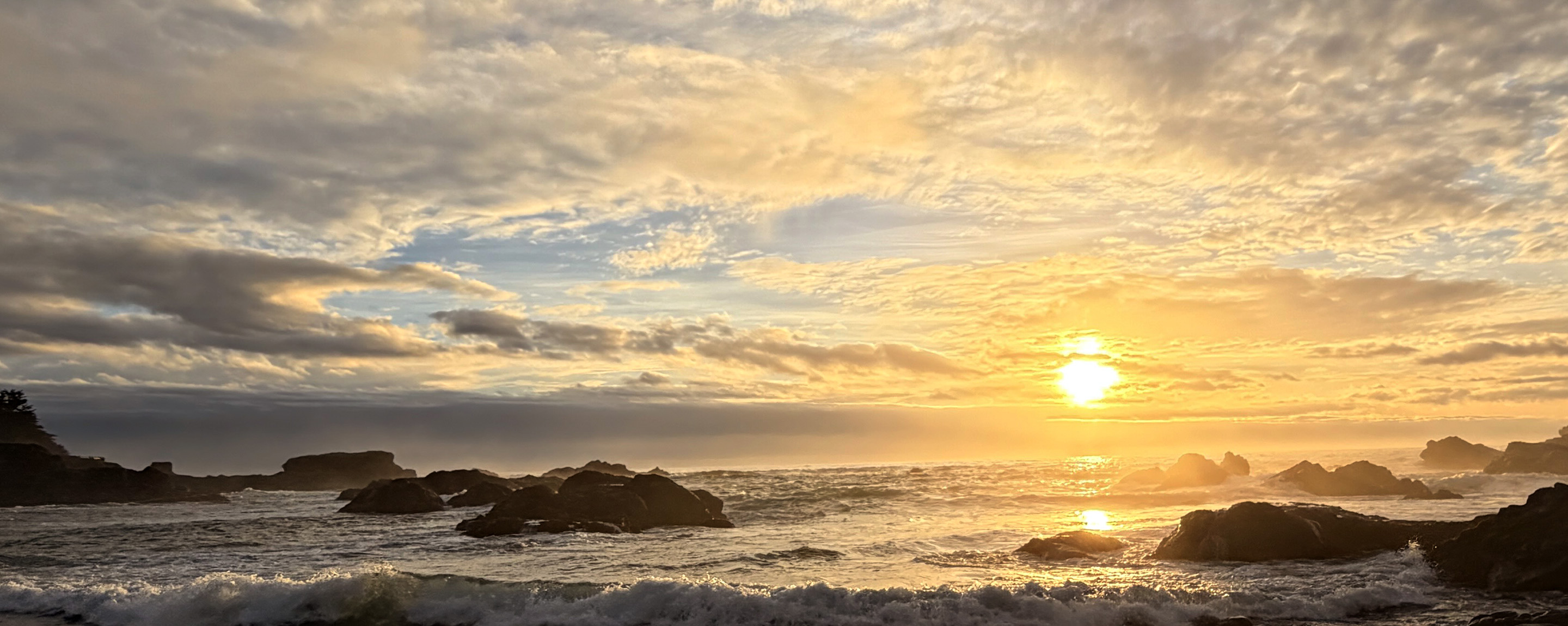 Golden sunset over the ocean with sun low on the horizon, soft clouds overhead, and rocks and surf along the shore